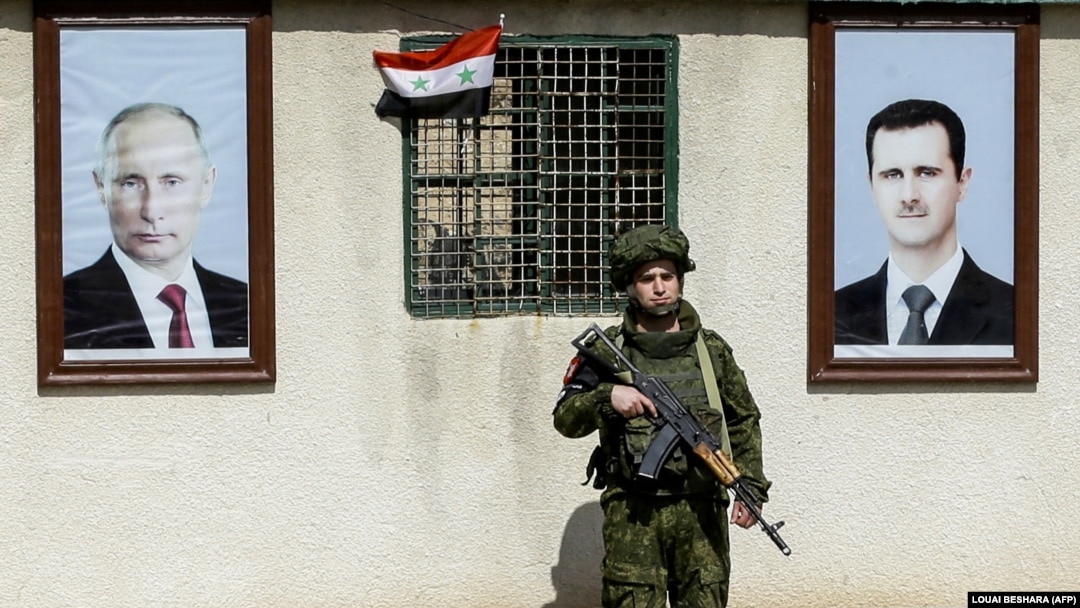 Російський військовий стоїть між портретами президента Сирії Башара Асада (праворуч) і президента Росії Володимира Путіна на блокпосту на околиці Дамаска, архівне фото, березень 2018 року